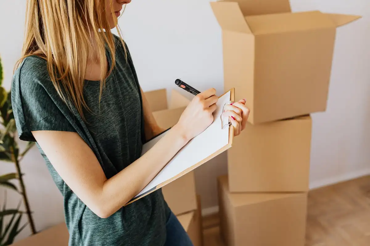 Mujer anota en un portapapeles junto a una pila de cajas de cartón mientras prepara el inventario de una mudanza