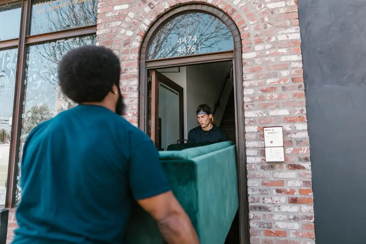Operario de espaldas carga un sofá verde para entrar a un edificio de ladrillo con puerta en arco durante una mudanza